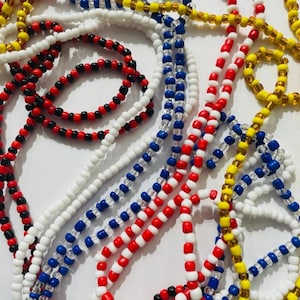 Può includere: Una collezione di collane di perline in vari colori, tra cui rosso, bianco, blu, giallo e nero. Le perline sono piccole e infilate insieme, creando un'esposizione strutturata e colorata. Le collane sono probabilmente per scopi decorativi o spirituali.