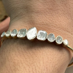 Könnte beinhalten: Ein goldfarbenes Armband mit einer Reihe unregelmäßig geformter, klarer Edelsteine. Jeder Edelstein ist in einer goldenen Lünette gefasst. Das Armband hat ein geschwungenes Design mit einem schlanken Goldband. Die Edelsteine sind unterschiedlich groß und geformt.