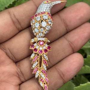 Può includere: Una spilla a forma di colibrì in metallo dorato, con testa e corpo incastonati di pietre bianche trasparenti. La coda è composta da piume a ventaglio sovrapposte, decorate con file alternate di pietre rosa vivace e bianche trasparenti.