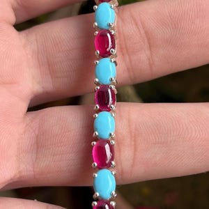 May include: A silver bracelet with alternating oval turquoise and ruby gemstones. The gemstones are set in a row, creating a vibrant contrast of colours. The bracelet is a piece of jewellery.