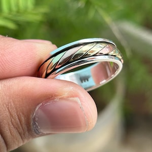 May include: A silver and gold spinning ring with a braided design. The ring is worn on a finger.