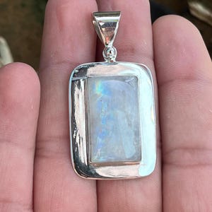 Könnte beinhalten: Ein silberner Anhänger mit einem rechteckigen Mondstein, der in einen silbernen Rahmen gefasst ist. Der Mondstein hat eine milchig weiße Farbe mit schillernden blauen und grünen Blitzen.