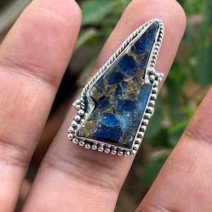 May include: A silver ring with a blue and brown gemstone set in a triangular shape. The ring has a beaded edge.