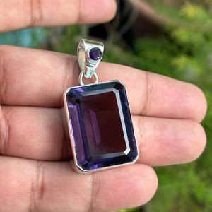 Pode incluir: Um pingente de prata com uma grande pedra de ametista retangular de cor púrpura escura. O pingente tem uma pedra de ametista redonda menor na parte superior.