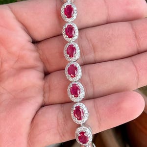 Könnte beinhalten: Ein silbernes Armband mit ovalen, tiefroten Rubinen, umgeben von kleinen, klaren Edelsteinen. Das Armband wird in einer Hand gehalten, um das Design und die Farbe des Schmucks hervorzuheben. Die Edelsteine sind in einem symmetrischen Muster angeordnet.