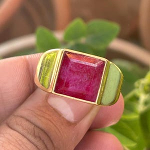 May include: A gold ring featuring a large, square, red gemstone set in the centre. The ring has a gold band with a green enamel accent on either side of the stone.