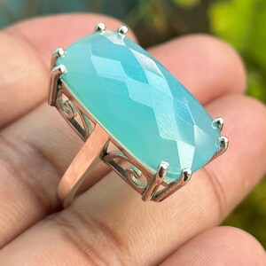 Puede incluir: Un anillo de plata con una gran piedra preciosa azul claro. La piedra preciosa está facetada y tiene una forma rectangular.
