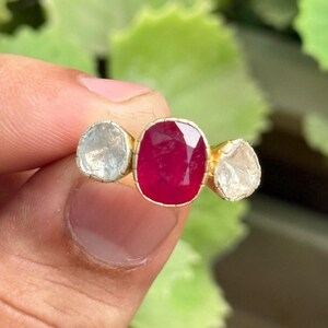 Puede incluir: Un anillo de oro con un rubí central de talla ovalada y dos piedras más pequeñas, de color claro y forma irregular. El anillo se muestra sobre un fondo verde borroso.