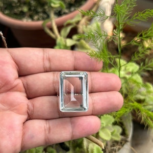 Puede incluir: Un anillo de plata con una piedra rectangular transparente. El anillo está en una mano, con follaje verde en el fondo.