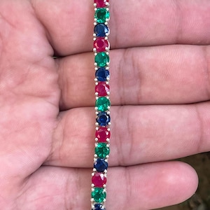 Puede incluir: Un brazalete de tenis de plata con piedras redondas alternas de color rojo, verde y azul.