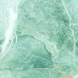 Könnte beinhalten: Nahaufnahme einer Natursteinoberfläche mit marmoriertem Aussehen. Der Stein weist eine Mischung aus Grün- und Weißtönen mit komplizierten Maserungsmustern auf. Die Textur ist rau und uneben und erzeugt eine einzigartige Ästhetik.