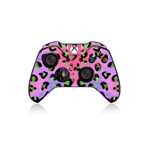 Puede incluir: Un mando de Xbox rosa y negro con un diseño de estampado de leopardo. El mando tiene una placa frontal negra con botones verdes, amarillos y azules.