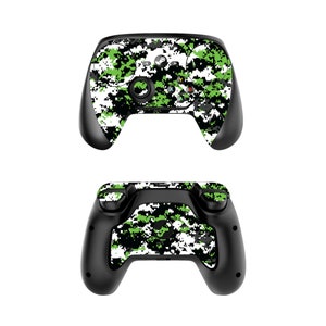Puede incluir: Un mando de videojuego con estampado de camuflaje negro y verde. El mando tiene una base negra y un estampado de camuflaje verde y blanco en la parte superior y los laterales. El mando tiene un botón circular en el centro y dos botones más pequeños a cada lado.