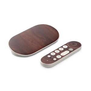 Op de afbeelding: Een set elektronische apparaten met een houtnerf afwerking. De set bevat een rechthoekige basis en een afstandsbediening met witte knoppen. De afstandsbediening heeft knoppen voor YouTube, Netflix en andere functies. De apparaten hebben een lichtgekleurde rand.