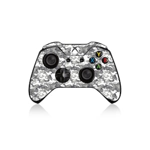 Może przedstawiać: Kontroler Xbox z szarym, białym i czarnym wzorem kamuflażu. Kontroler ma czarne joysticki, d-pad i kolorowe przyciski akcji. Logo Xbox znajduje się w górnej części.