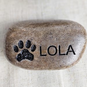 May include: A light brown stone with a black paw print and the name "LOLA" engraved on it.
