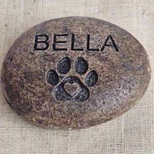 May include: A brown, round, engraved stone with the name "BELLA" and a paw print with a heart inside.