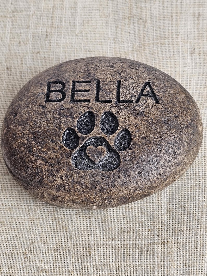 May include: A brown, round, engraved stone with the name "BELLA" and a paw print with a heart inside.