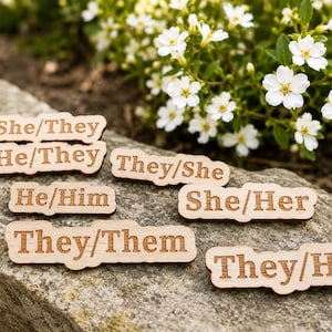 May include: A collection of six light-coloured wooden pronoun pins with laser-engraved text. The pins display the following pronoun sets: She/They, He/They, They/She, He/Him, She/Her, They/Them, and They/He, each featuring a smooth, natural wood finish.