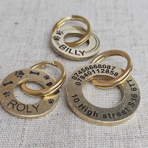 May include: Three gold-coloured metal dog tags with engraved text. The first tag reads "ROLY" and has paw prints and a bone design. The second tag reads "BILLY" and has paw prints. The third tag has a phone number, address, and the text "10 High Street 36 6TT".