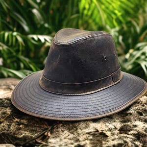 May include: A dark brown leather hat with a wide brim and a band around the crown. The hat has a distressed look, with visible wear and tear. The hat is sitting on a rock, with a blurred green background of foliage.