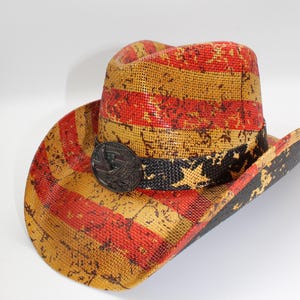 May include: A straw cowboy hat with a distressed American flag design in red, yellow, and black. The hat features a black band with white stars and a metal emblem. The brim is wide and the crown is rounded.