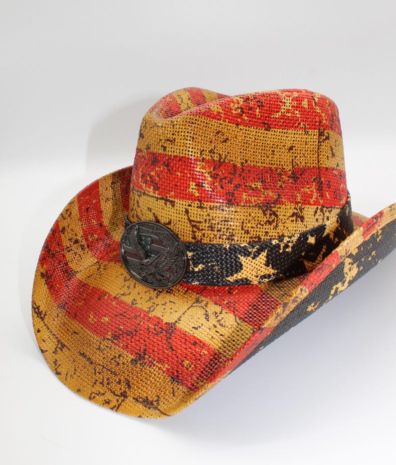 May include: A straw cowboy hat with a distressed American flag design in red, yellow, and black. The hat features a black band with white stars and a metal emblem. The brim is wide and the crown is rounded.