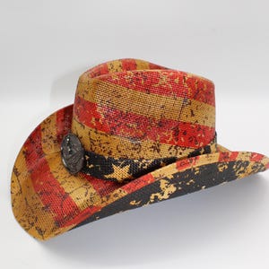 May include: A distressed-look cowboy hat with red and tan stripes, a black band, and a decorative metal emblem. The hat has a wide brim and a structured crown, suitable for sun protection and fashion.