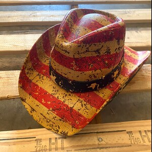 May include: A straw cowboy hat with a distressed American flag design. The hat features red and yellow stripes with a black band around the crown. The hat is resting on a wooden pallet.