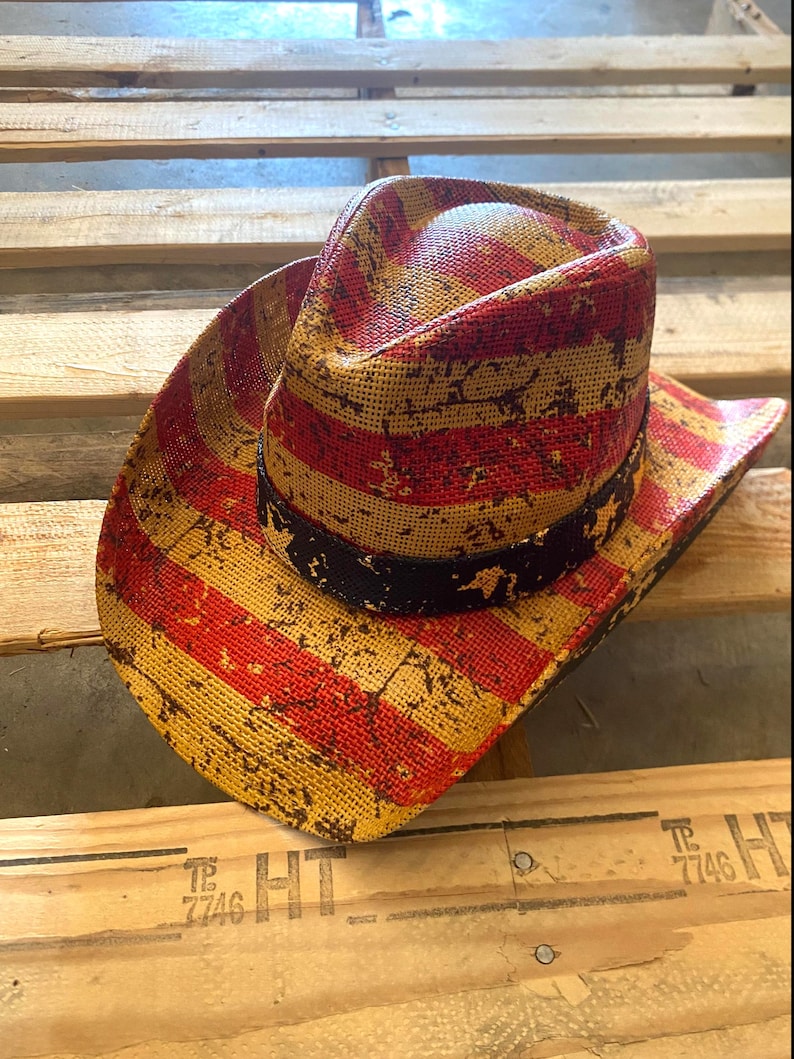 May include: A straw cowboy hat with a distressed American flag design. The hat features red and yellow stripes with a black band around the crown. The hat is resting on a wooden pallet.