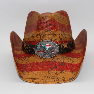 May include: A straw cowboy hat with a distressed American flag design in red and tan. The hat features a black band with a silver buckle depicting an eagle and the American flag.
