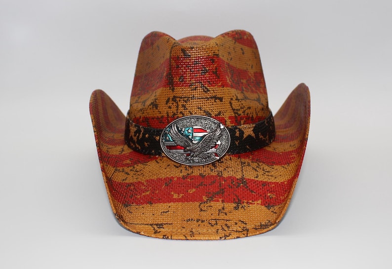 May include: A straw cowboy hat with a distressed American flag design in red and tan. The hat features a black band with a silver buckle depicting an eagle and the American flag.
