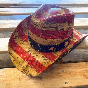 May include: A straw cowboy hat with a red, yellow, and black American flag design. The hat features a distressed pattern and a black band around the crown. The hat is resting on a wooden surface.