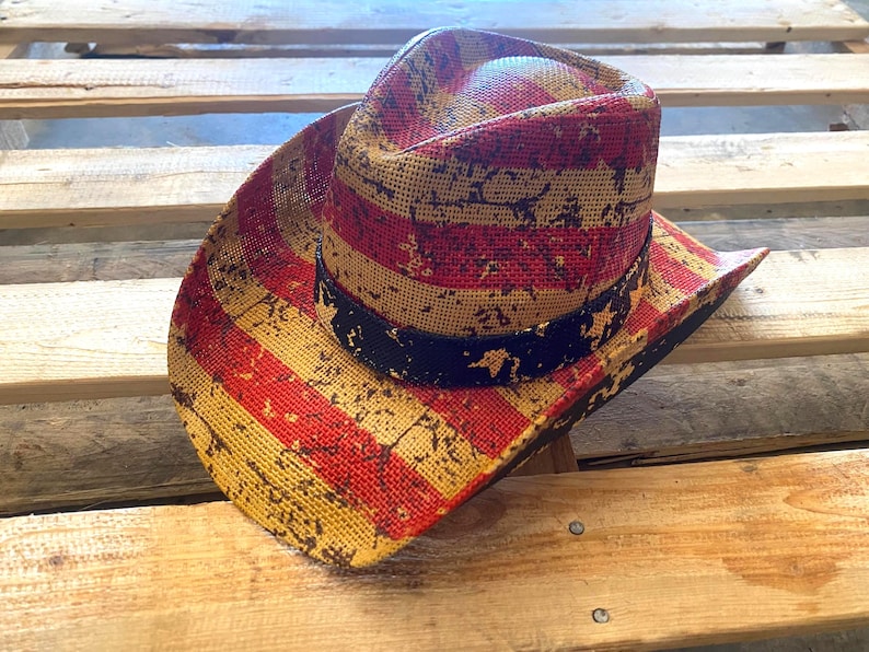 May include: A straw cowboy hat with a red, yellow, and black American flag design. The hat features a distressed pattern and a black band around the crown. The hat is resting on a wooden surface.