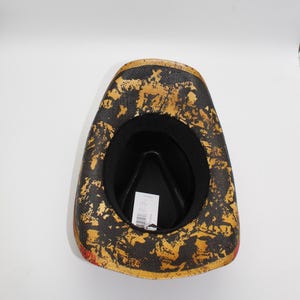 May include: A cowboy hat with a black interior and a textured exterior featuring gold and black patterns. The hat has a red trim along the edge and a tag inside.