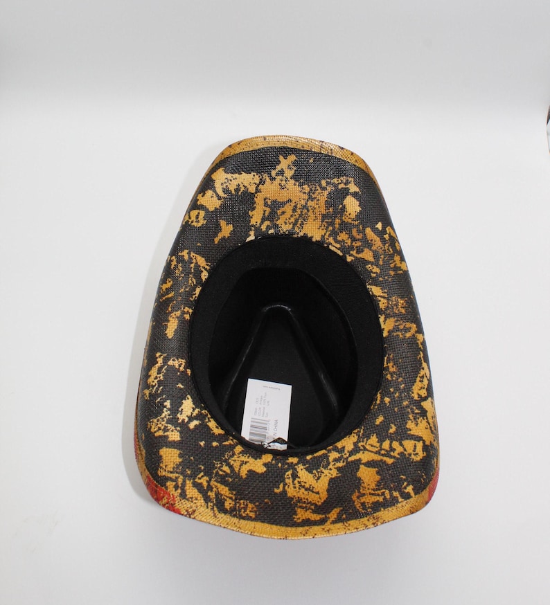 May include: A cowboy hat with a black interior and a textured exterior featuring gold and black patterns. The hat has a red trim along the edge and a tag inside.