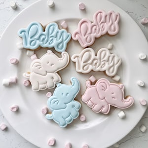 May include: Six decorated sugar cookies on a white plate. Three cookies are decorated with the word "Baby" in blue icing. Three cookies are decorated with a pink elephant.