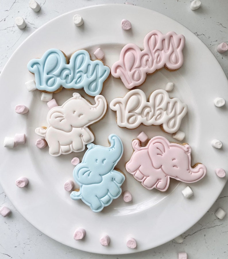 May include: Six decorated sugar cookies on a white plate. Three cookies are decorated with the word "Baby" in blue icing. Three cookies are decorated with a pink elephant.