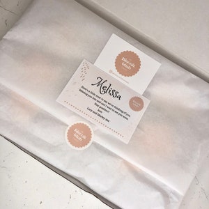 May include: A white gift box with a white tissue paper liner. A card with the text "Melissa" and "Here's a little treat to say we're thinking of you. Missing you lots and can't wait to see you soon. Stay positive!! Love, Lucy and Marley xxx" is on top of the tissue paper. The card has a brown circle with the text "The Biscuit Club" in white.