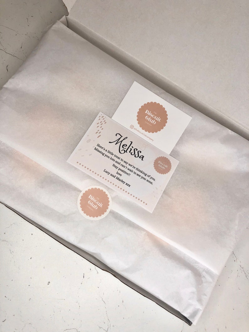 May include: A white gift box with a white tissue paper liner. A card with the text "Melissa" and "Here's a little treat to say we're thinking of you. Missing you lots and can't wait to see you soon. Stay positive!! Love, Lucy and Marley xxx" is on top of the tissue paper. The card has a brown circle with the text "The Biscuit Club" in white.