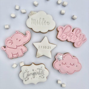 May include: A set of six decorated sugar cookies for a baby shower. The cookies are decorated with pink and white icing and feature an elephant, a cloud, a star, and the words "Baby", "Congrats", and "Millie".