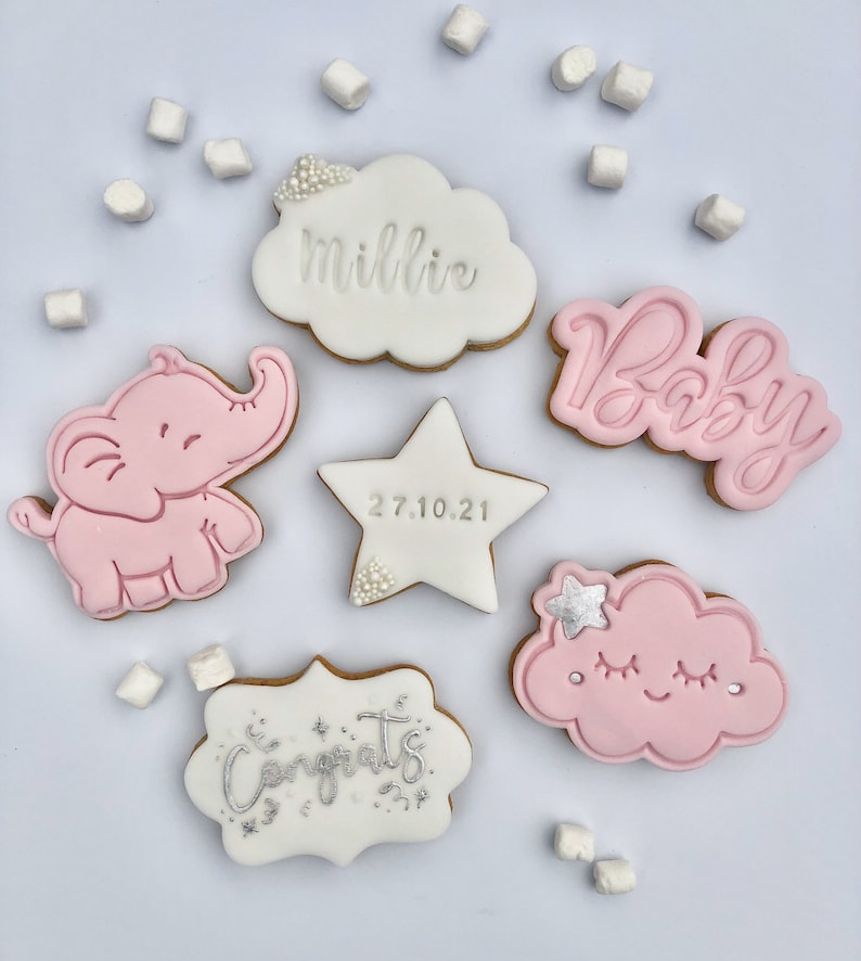 May include: A set of six decorated sugar cookies for a baby shower. The cookies are decorated with pink and white icing and feature an elephant, a cloud, a star, and the words "Baby", "Congrats", and "Millie".