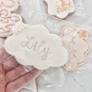 May include: A white cloud-shaped cookie with the name "Lily" written in cursive on it. The cookie is decorated with small white flowers and is packaged in a clear plastic bag.