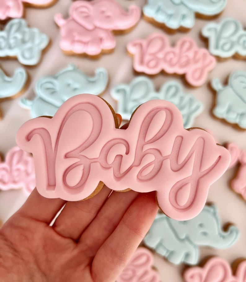 May include: Pink and blue sugar cookies decorated with elephant and baby designs. The cookies are decorated with royal icing and the word "Baby" is written on one of the cookies.