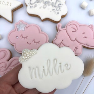 May include: A set of decorated sugar cookies with pink and white icing. One cookie is shaped like a cloud and says "Millie" on it. Another cookie is shaped like an elephant and is pink. A third cookie is shaped like a cloud with a pink and white star on it. The cookies are on a white surface.