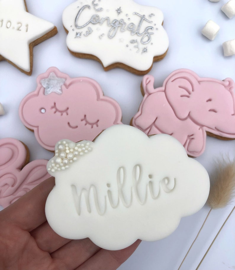 May include: A set of decorated sugar cookies with pink and white icing. One cookie is shaped like a cloud and says "Millie" on it. Another cookie is shaped like an elephant and is pink. A third cookie is shaped like a cloud with a pink and white star on it. The cookies are on a white surface.