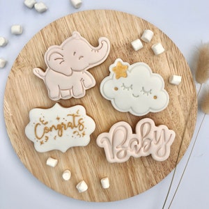 May include: Four decorated sugar cookies on a wooden surface. The cookies are decorated with pink and white icing and gold lettering. One cookie is shaped like an elephant, another is shaped like a cloud, and the other two are shaped like banners. The banners say "Congrats" and "Baby".