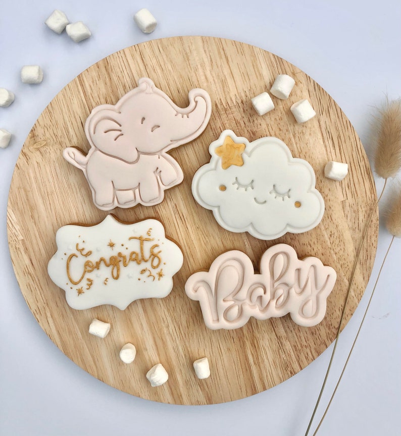 May include: Four decorated sugar cookies on a wooden surface. The cookies are decorated with pink and white icing and gold lettering. One cookie is shaped like an elephant, another is shaped like a cloud, and the other two are shaped like banners. The banners say "Congrats" and "Baby".