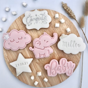 May include: A set of six decorated sugar cookies on a wooden board. The cookies are decorated with pink and white icing and feature a cloud, an elephant, a star, a banner that says "Congrats", a cloud that says "Millie", and a cookie that says "Baby".