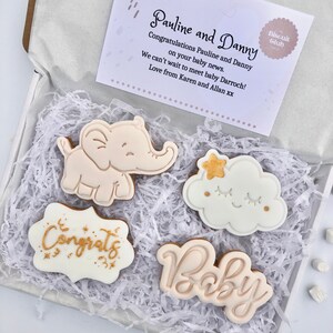 May include: A gift box filled with white shredded paper and four decorated cookies. The cookies are decorated with gold and white icing and feature a baby elephant, a cloud with a star, the word "Congrats" and the word "Baby". The box also includes a card with the text "Pauline and Danny Congratulations Pauline and Danny on your baby news. We can't wait to meet baby Darroch! Love from Karen and Allan xx" and a small sticker that says "Biscuit Club".